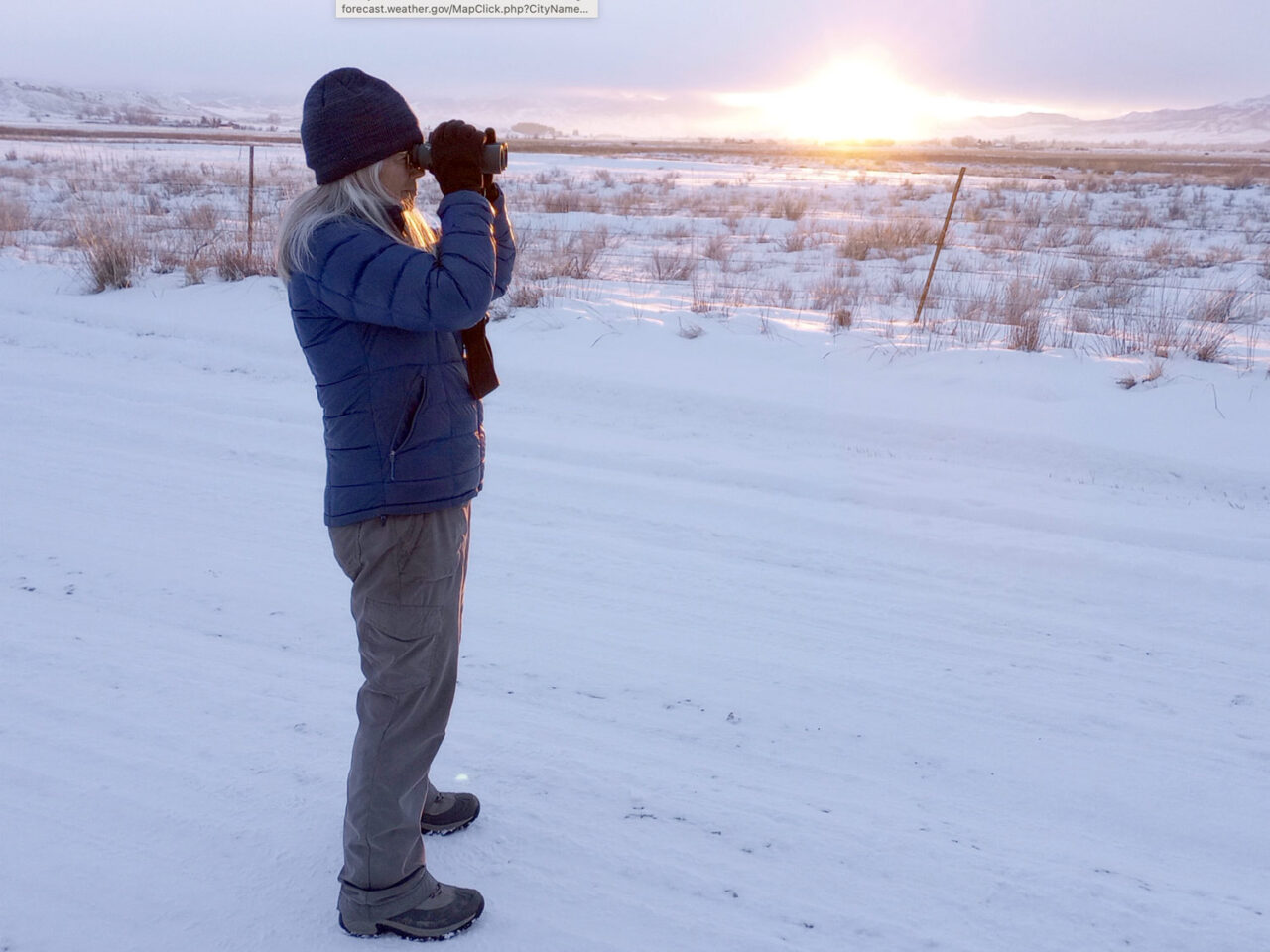 A person looks through binoculars at snow-covered prairie.