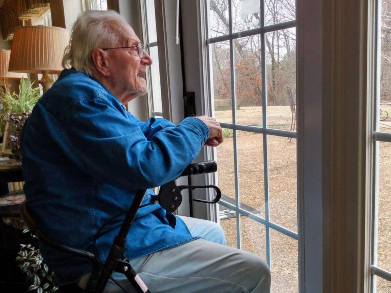 An elderly person rests on a walker and looks outside through windows.
