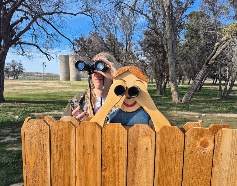 A person using binoculars to birdwatch while standing next to a wooden cutout of a person with binoculars.