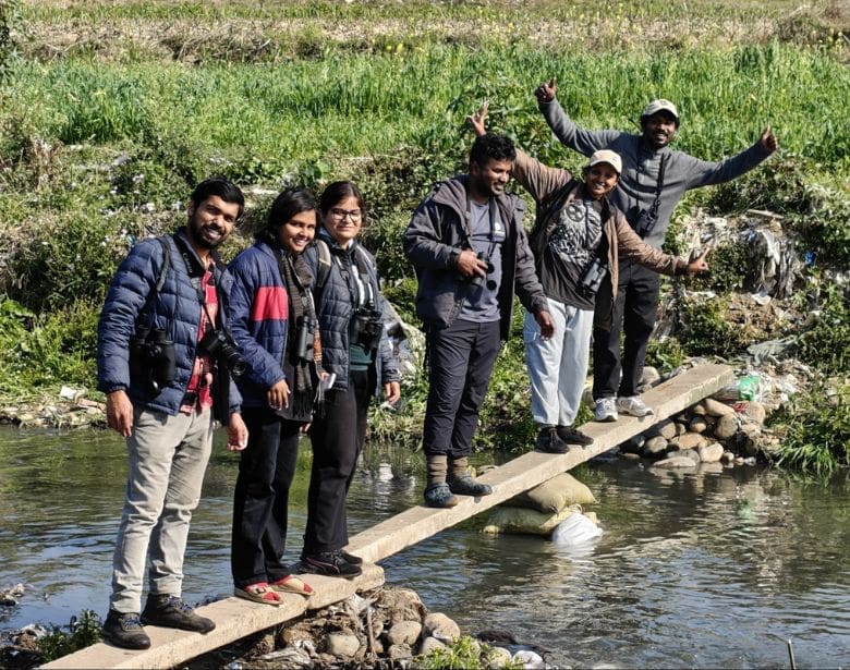 Group of smiling people with binoculars, standing on a wooden plank. The wooden plank is over a small body of water.