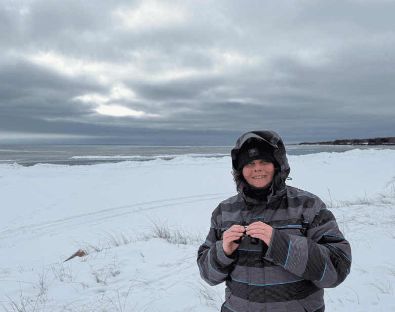 A person smiling and holding a pair of binoculars in front of a body of water.