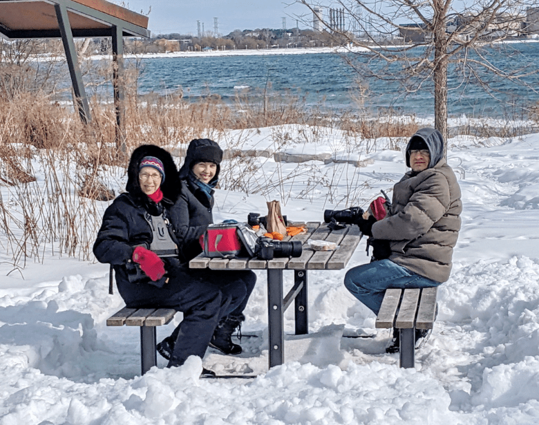Three smiling people sitting at an outdoor table in front of a body of water.