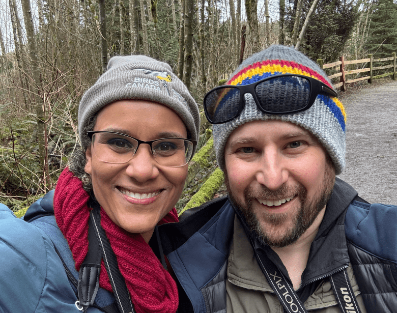 Two smiling people in a wooded area.