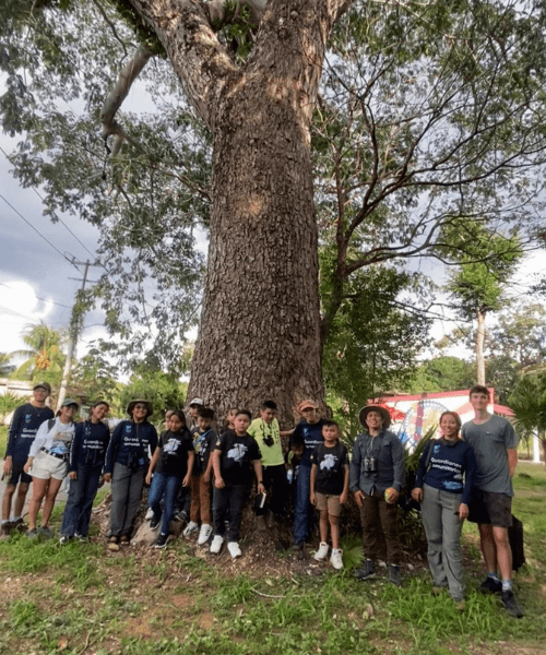 A group of smiling people with birdwatching equipment standing in front of a tree.