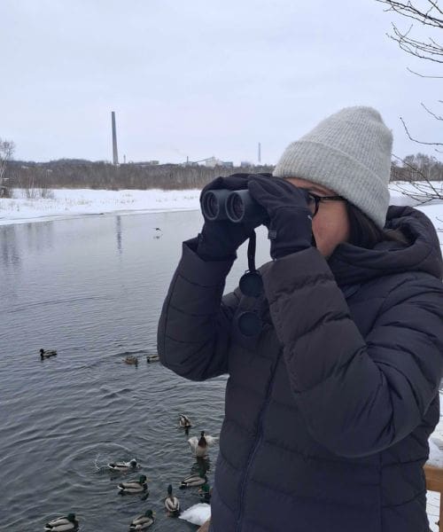 A person birdwatching with binoculars in front of a body of water.