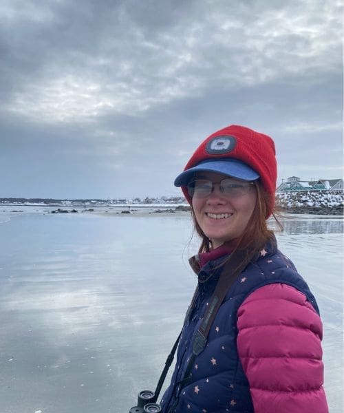 A person smiling while holding binoculars in front of a body of water.